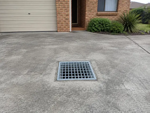 Square metal gully grate set in a dry concrete driveway beside a brick house in Swansea