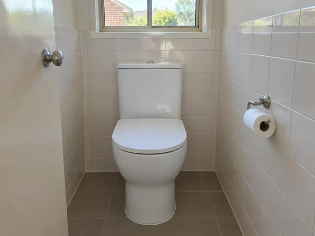 White toilet in a tiled small bathroom with window and toilet roll holder in Murrays Beach
